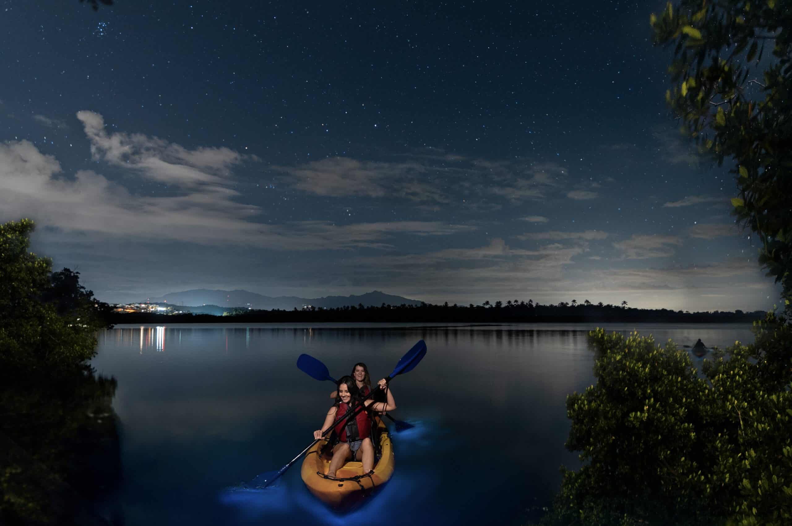 A dark cloudy night with stars showing and two people in a yellow kayak with glowing waters underneath them.